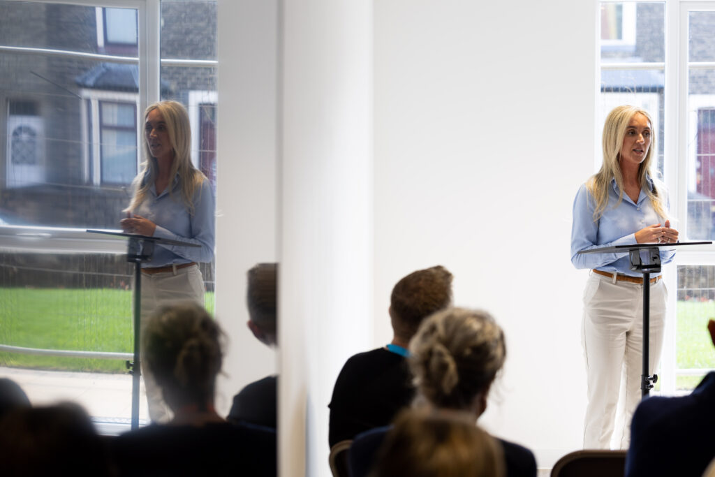 A person speaking at a podium in a bright room, with an audience seated in front.