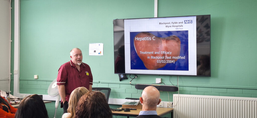 A speaker presents to colleagues in a meeting room beside a screen displaying information about Hepatitis C treatment and efficacy in Blackpool.